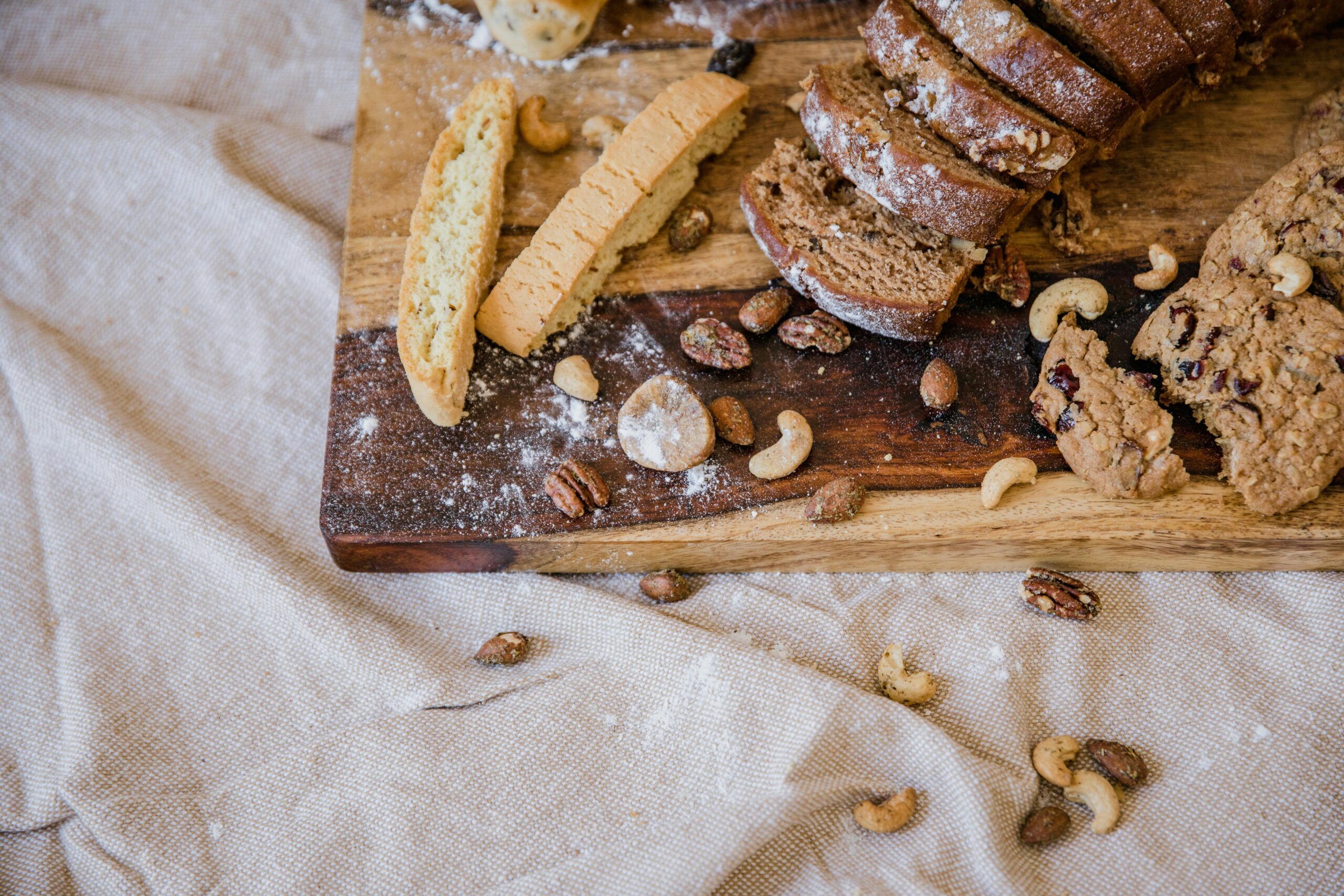 Rustic assortment of sliced bread, cookies, and nuts on a wooden board over textile cloth.